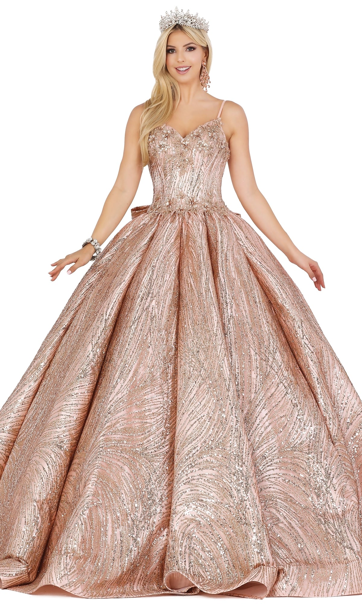 A woman in the Quinceanera Dress 1447 by Dancing Queen, featuring sparkling lace embroidery in rose gold with thin straps and a full skirt, accessorized with a silver crown and bracelet, stands against a white background.